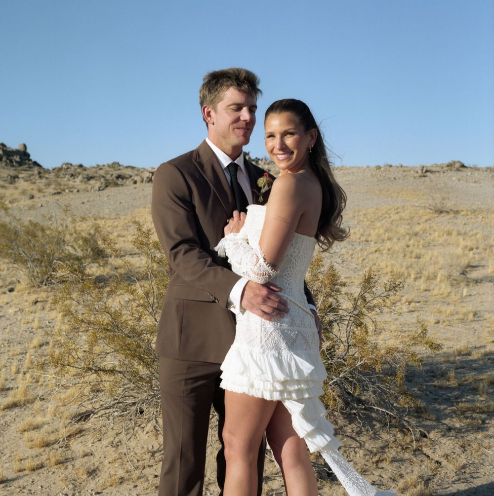 Couple in wedding attire embracing in a desert landscape, man in brown suit and woman in white dress.