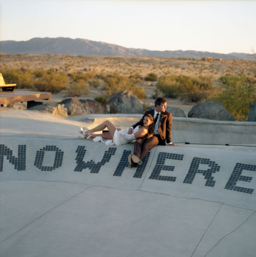 Couple sitting on Nowhere sign sculpture in desert landscape at golden hour.