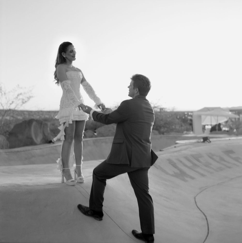 Black and white photo of groom kneeling holding bride's hand on a skate ramp outdoors.