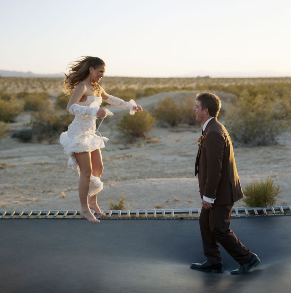 Bride in white mini dress jumps on trampoline while groom in brown suit watches in desert.