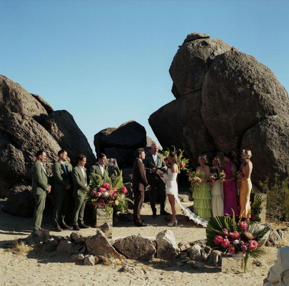Outdoor desert wedding ceremony with wedding party among large boulders under clear blue sky.