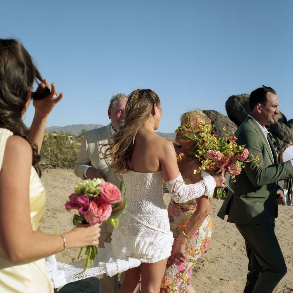Bride in white dress embraces guest while wedding party mingles outdoors in desert landscape.