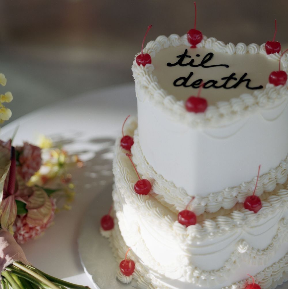 Two-tier white heart-shaped wedding cake decorated with red cherries and 'til death' inscription.