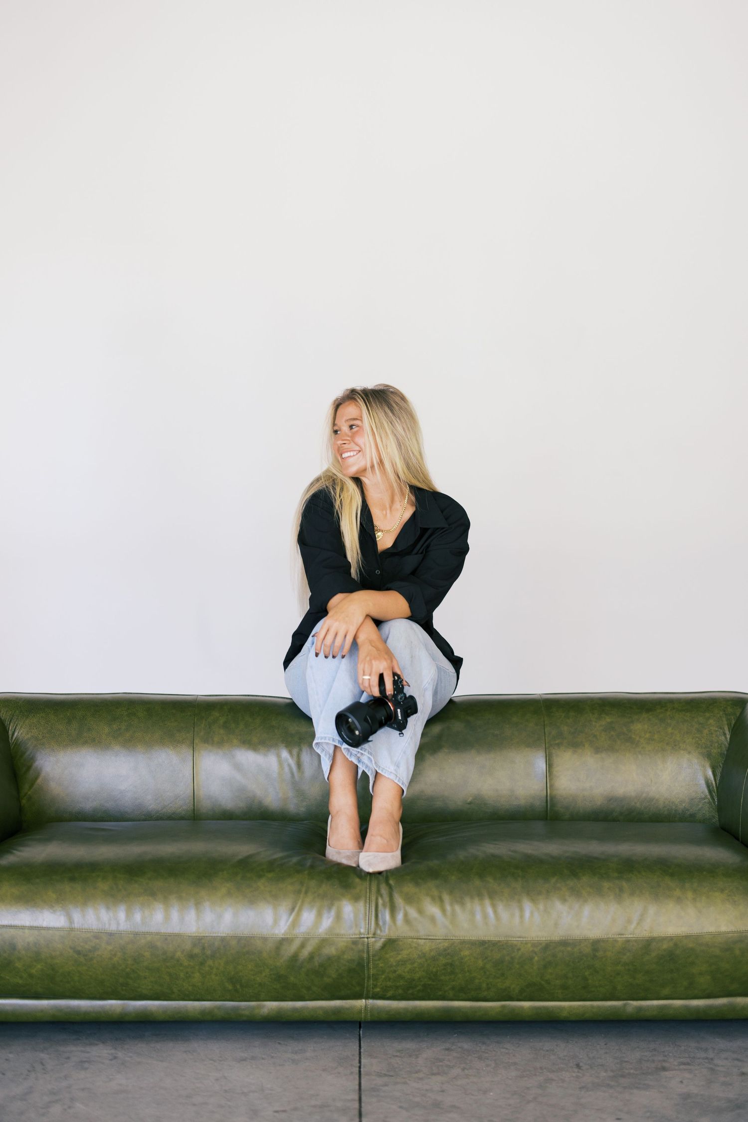 A woman in casual attire sits with camera on a vintage green leather couch against white wall.