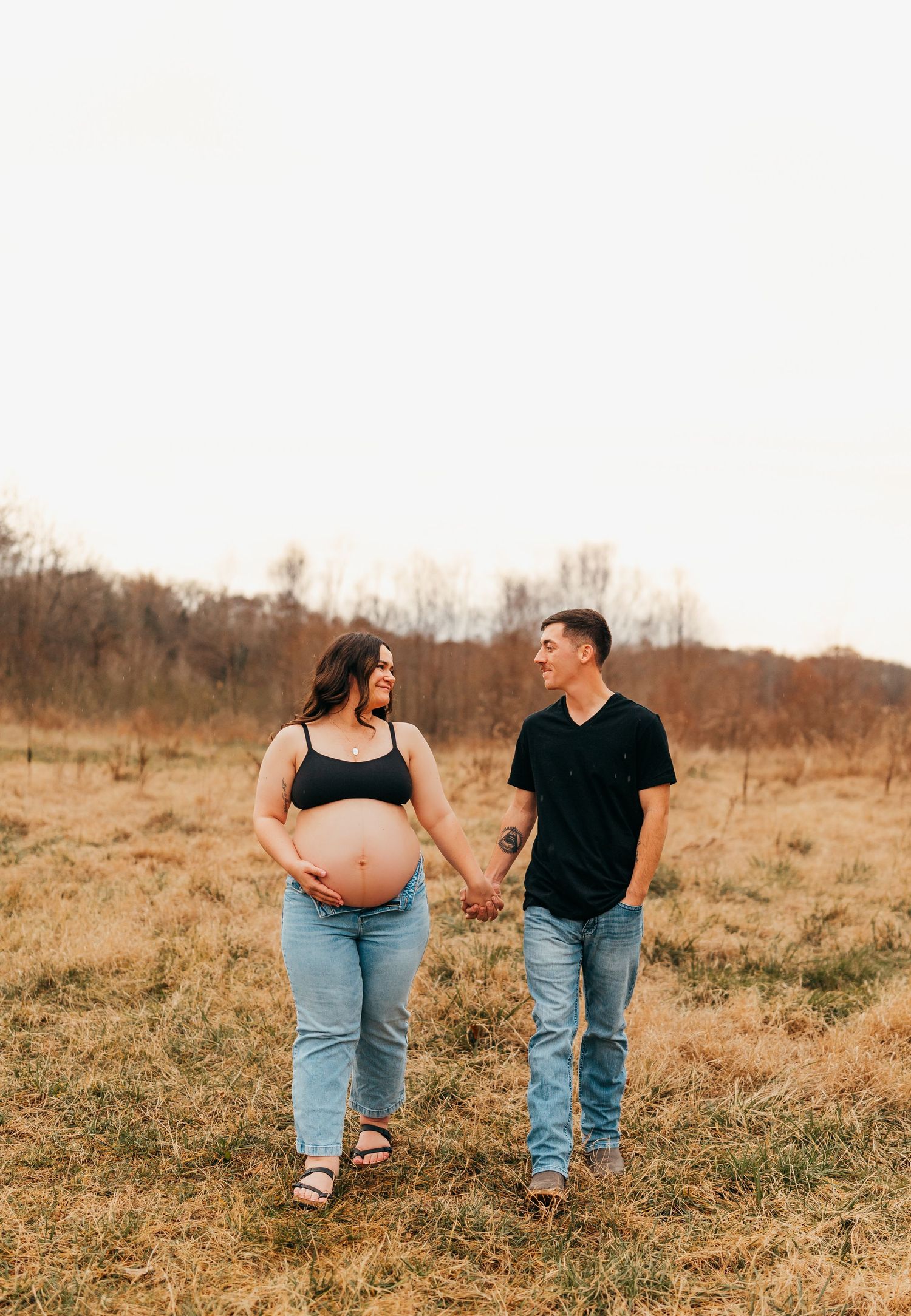 An expectant couple walks hand in hand through a rustic autumn field.