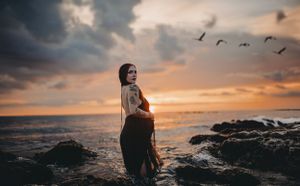 A silhouette poses against dramatic sunset sky with birds flying over ocean.