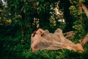 Woman in flowing sheer gold gown poses gracefully among lush green tropical trees in golden hour light.
