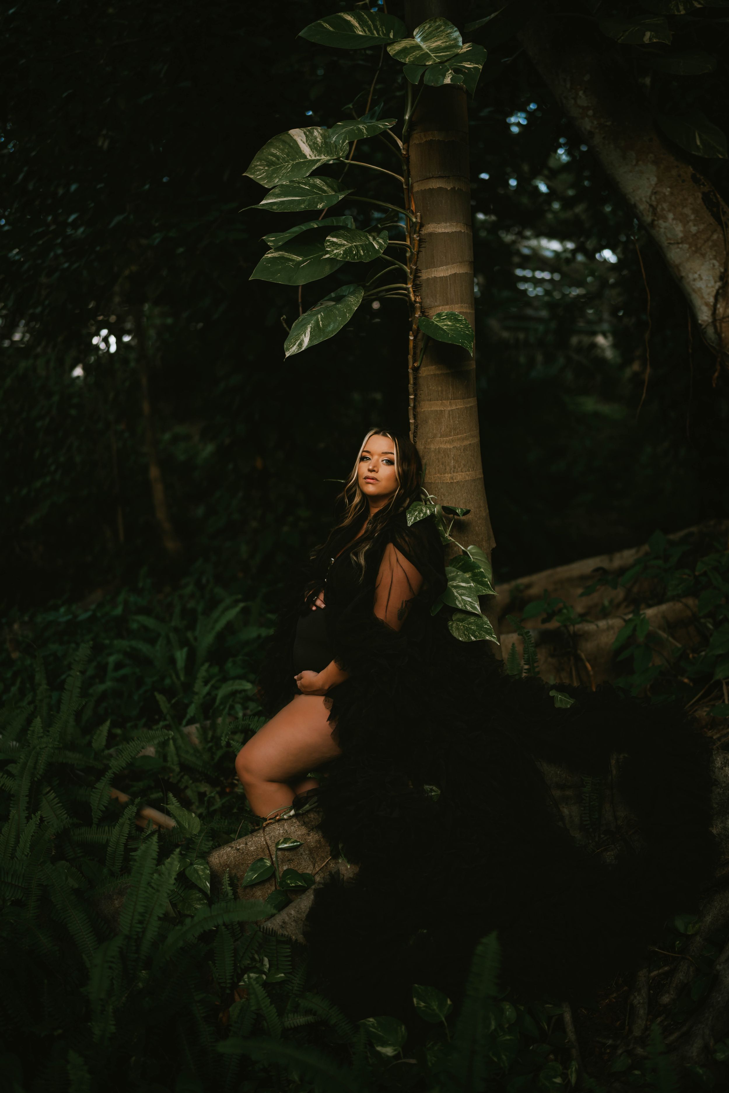 Woman in black dress and boots posing in a lush dark forest beside a tropical tree with large leaves.