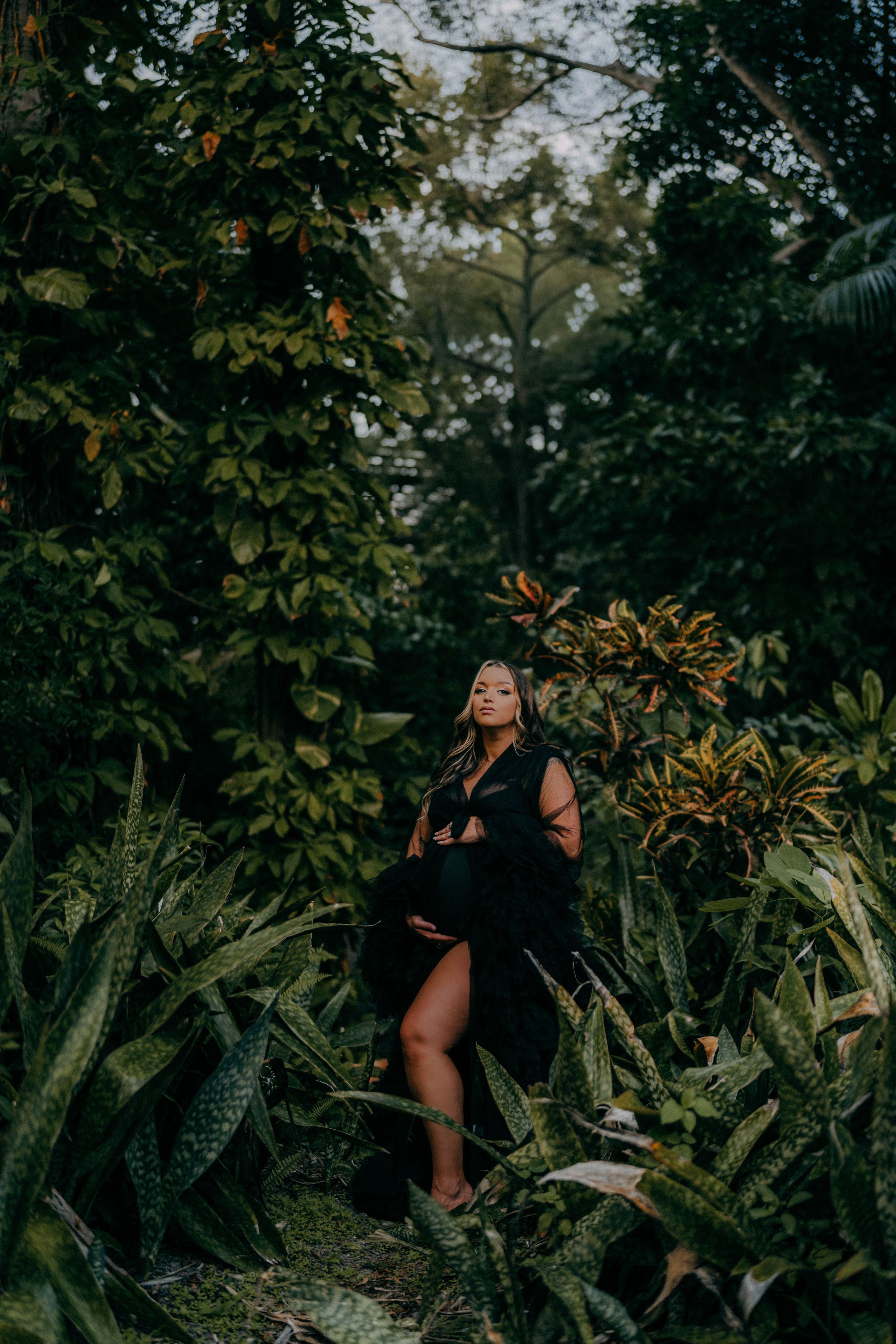 Woman in black dress posing gracefully among lush tropical greenery and exotic plants in a garden setting.