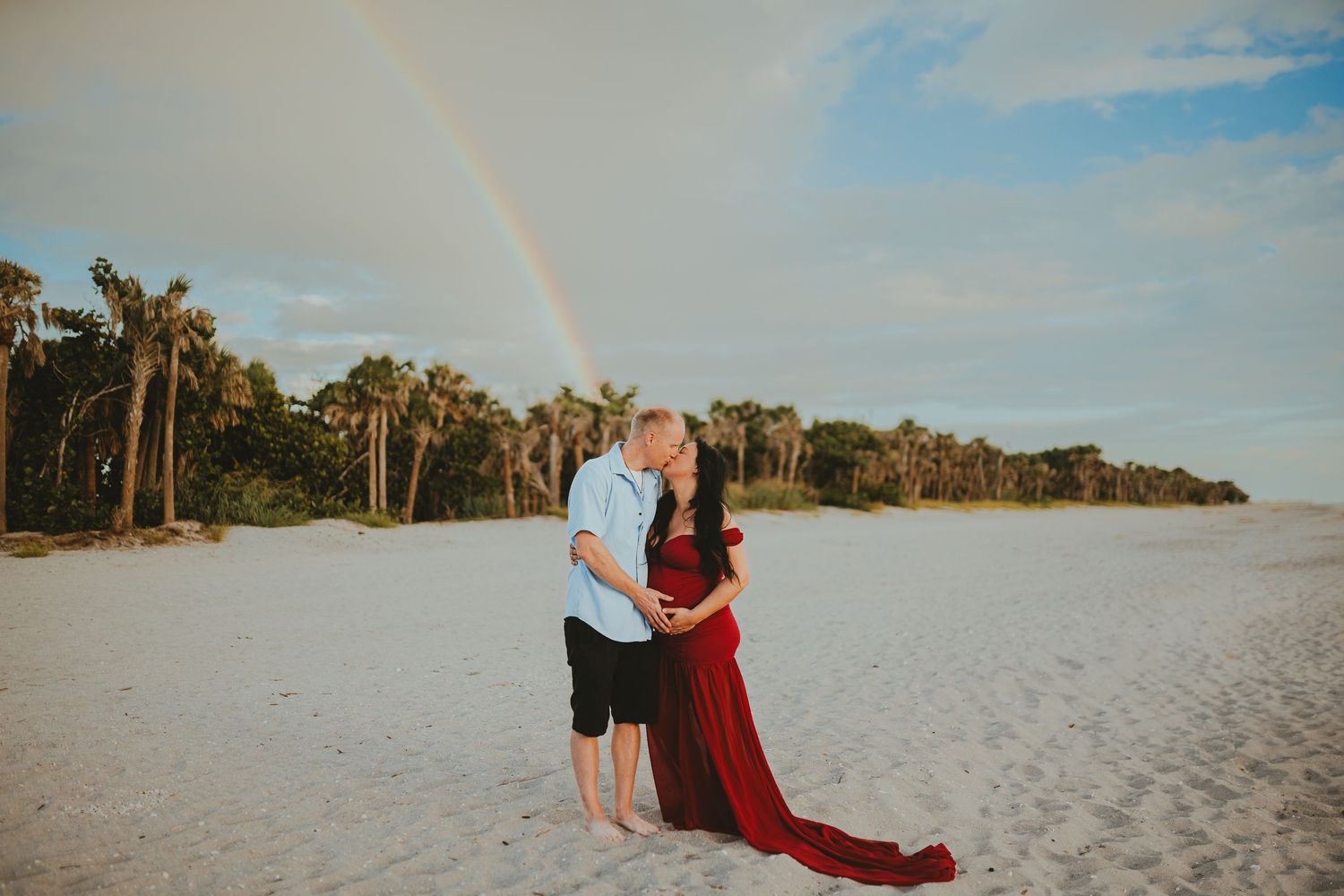 Pregnant couple kissing on white sand beach with rainbow and palm trees in background, maternity photo shoot.