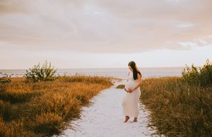 Pregnant woman in white dress standing on sandy beach path at golden sunset, cradling baby bump.