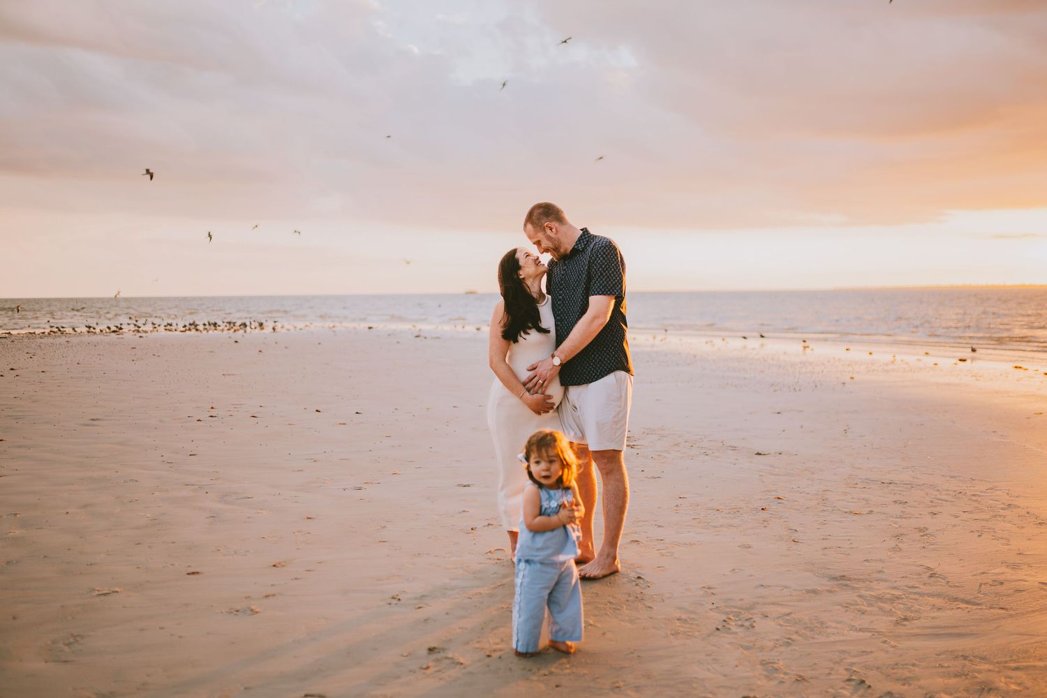 Couple holding hands on a sandy beach at sunset with a toddler standing in front of them.