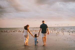 Family of three holding hands walking on beach at sunset with seagulls flying over ocean waves.