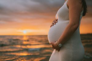 Pregnant woman in white dress cradling baby bump at golden sunset beach, ocean waves in background.