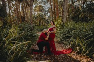 Couple in red outfits poses in lush green forest, man kneeling before pregnant woman in flowing gown.