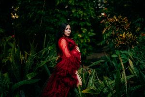 Pregnant woman in red tulle gown poses among lush tropical greenery in an elegant maternity photo.