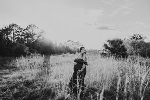 Black and white photo of a pregnant woman standing in tall grass fields during golden hour.