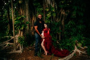 Couple poses in lush tropical forest, woman in flowing red maternity gown seated beside standing man in black shirt.