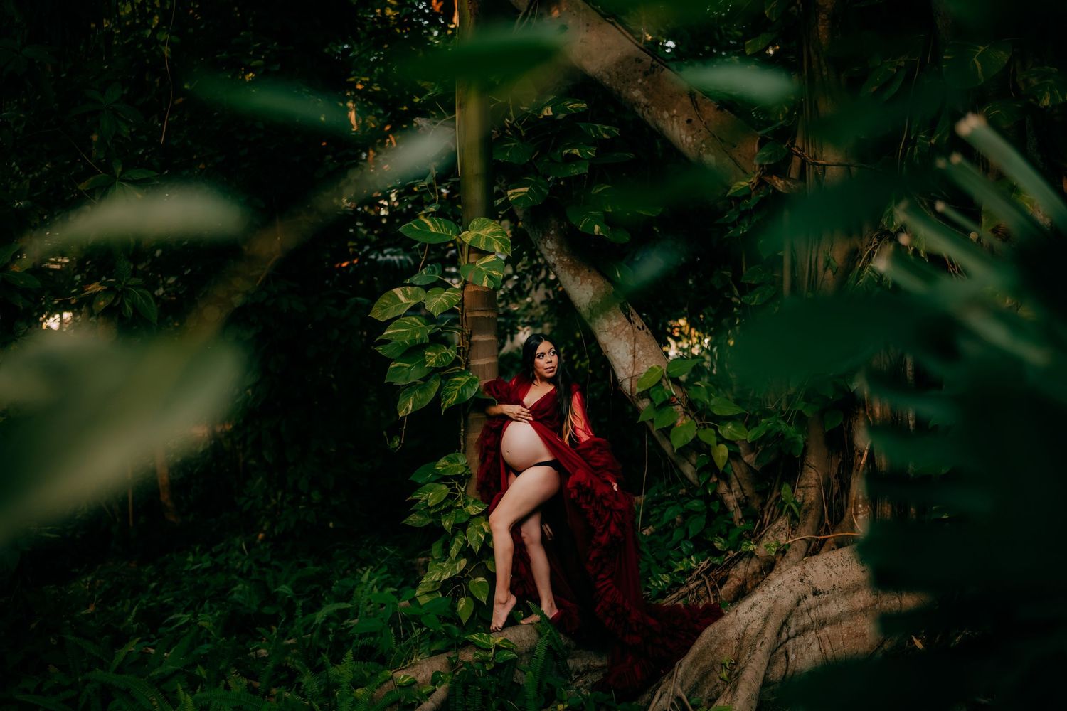 Pregnant woman in red gown poses among lush green tropical foliage, leaning against a tree in a serene outdoor setting.