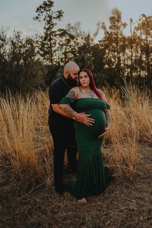 Couple maternity photo at golden hour, woman in green gown cradling baby bump, man embracing her in tall grass field.