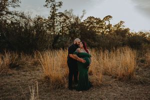 Couple embracing in golden field at sunset, woman in emerald green maternity gown, romantic outdoor pregnancy photo session.