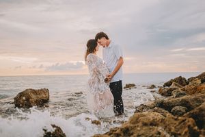 Romantic couple embracing on rocky beach at sunset with ocean waves crashing around them.