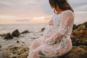 Pregnant woman in white lace gown sitting on rocks by the ocean at sunset, cradling her baby bump.