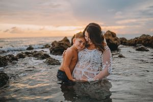 A mother and child embrace in ocean waters at sunset, with rocky shores and colorful sky behind them.