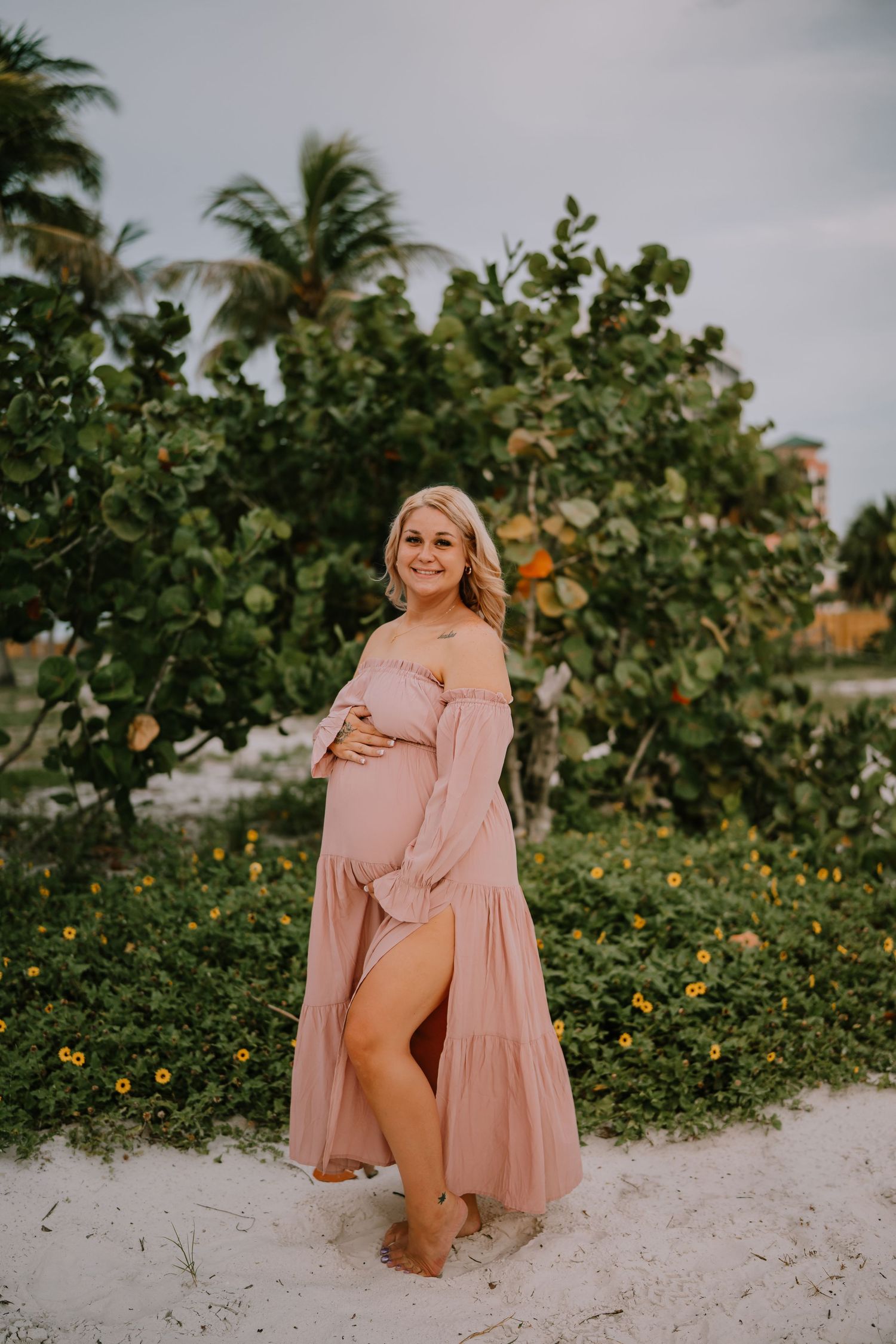 Pregnant woman in pink off-shoulder maxi dress poses on sandy beach with tropical greenery backdrop.