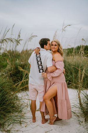 Couple embracing on beach dunes holding ultrasound photos, woman in pink off-shoulder maxi dress, pregnancy announcement photo.