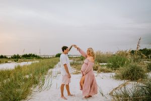 Pregnant couple dancing on white sand beach at sunset, woman in pink maxi dress, man in white shirt and shorts.