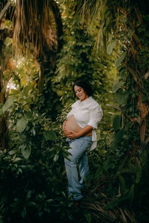 Pregnant woman in white shirt and jeans cradling her baby bump surrounded by lush tropical greenery.