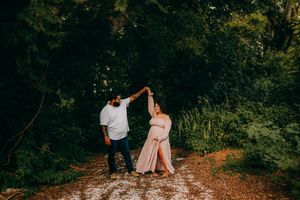 Couple dancing outdoors on a forest path, woman in pink maternity gown, man in white shirt, surrounded by lush green trees.