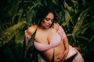 A plus-size woman in a pink lace bralette poses gracefully among lush tropical green leaves.