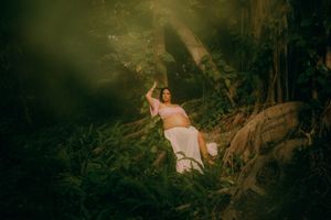 Pregnant woman in pink top and white skirt sitting in a lush green forest with dramatic sunlight filtering through trees.