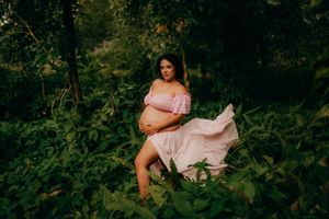 Pregnant woman in pink off-shoulder dress posing outdoors surrounded by lush green foliage during maternity photo shoot.