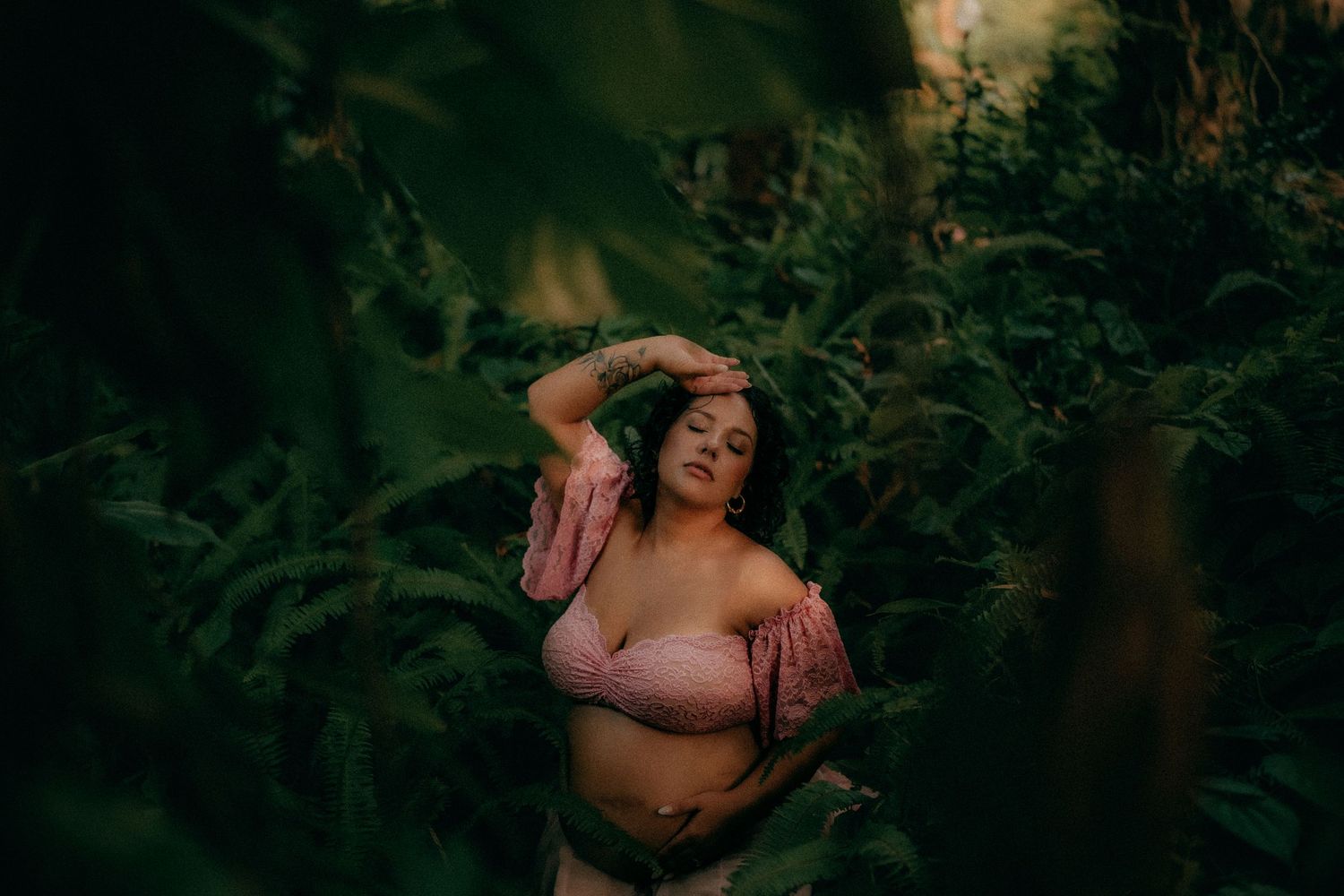 Pregnant woman in pink off-shoulder top poses among lush green ferns in a moody outdoor maternity photo.