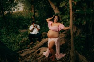Pregnant woman in pink off-shoulder gown poses by tree roots in lush forest during maternity photoshoot.