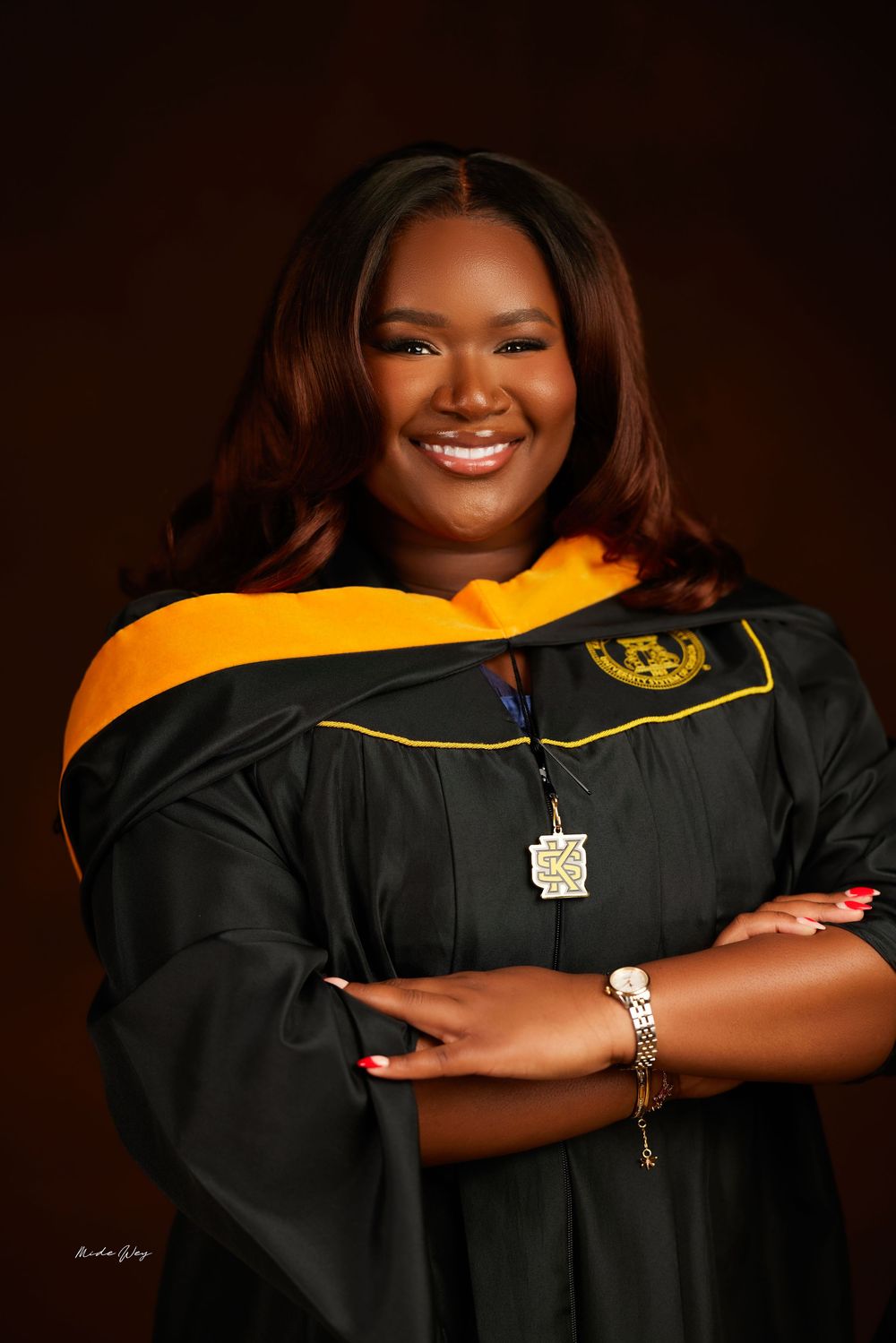 A graduate in academic regalia wearing a black graduation gown with gold trim and a necklace poses for a portrait.