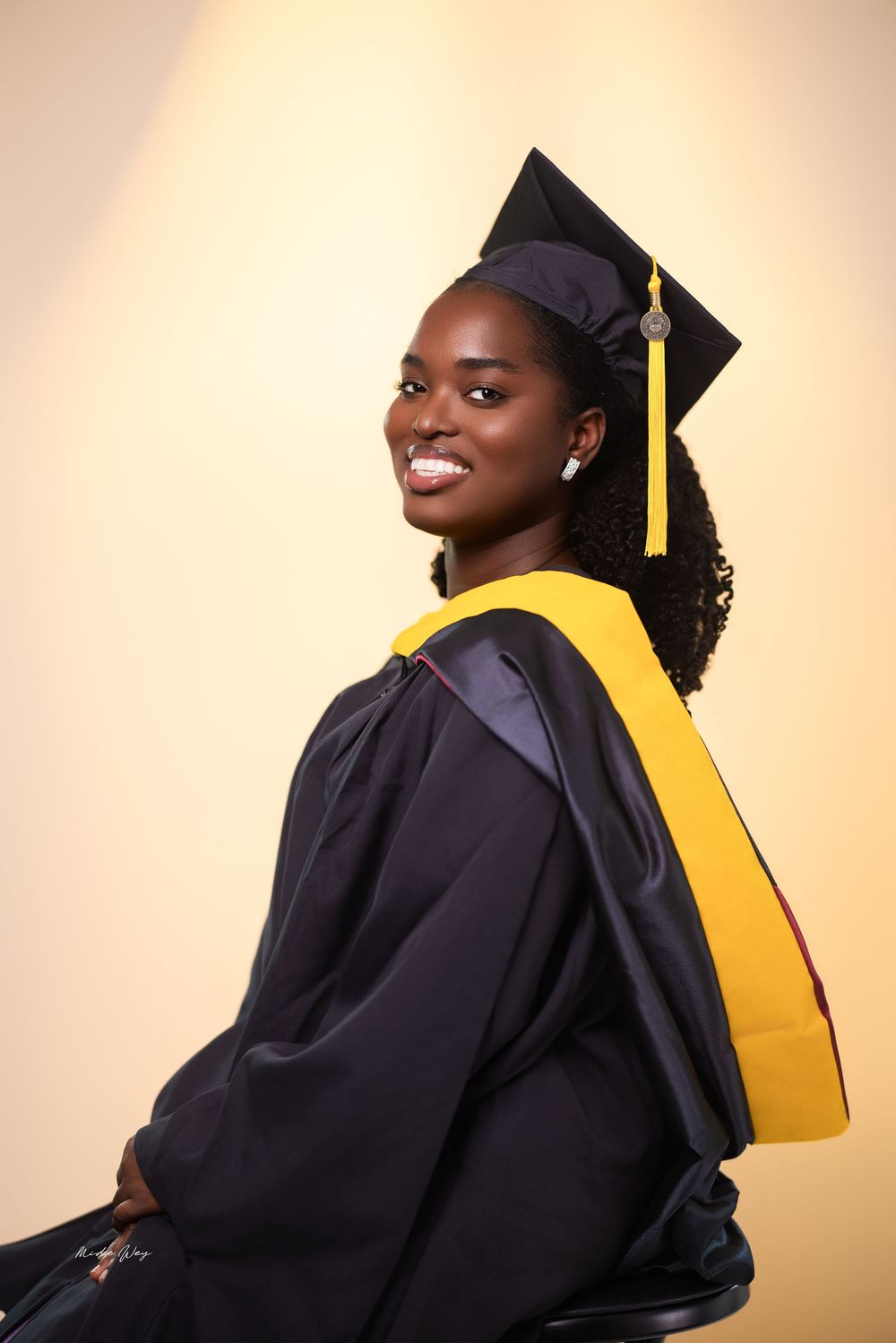 Graduate wearing black cap and gown poses against beige backdrop in professional portrait series.
