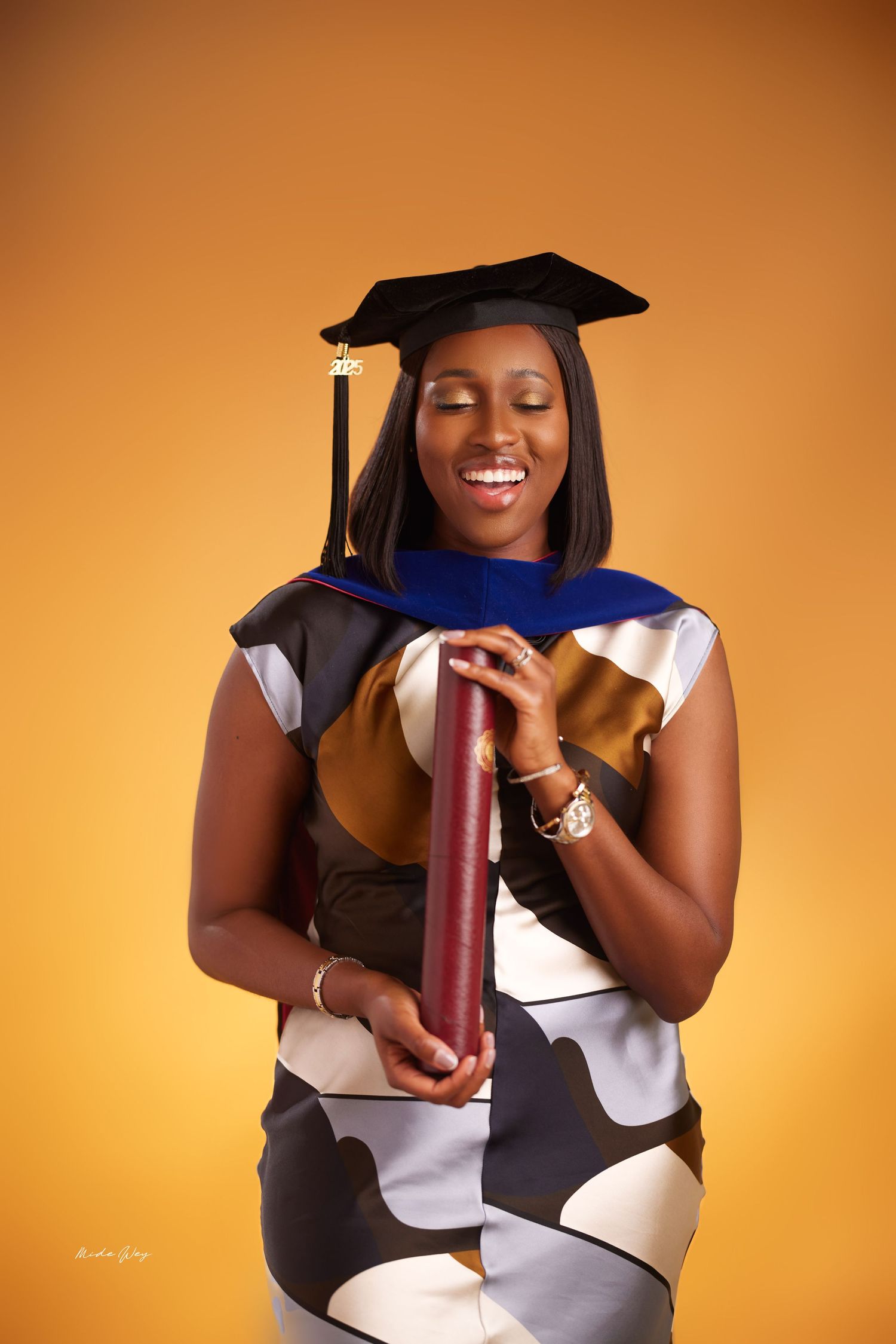 Graduate in academic regalia poses with diploma against warm orange background.