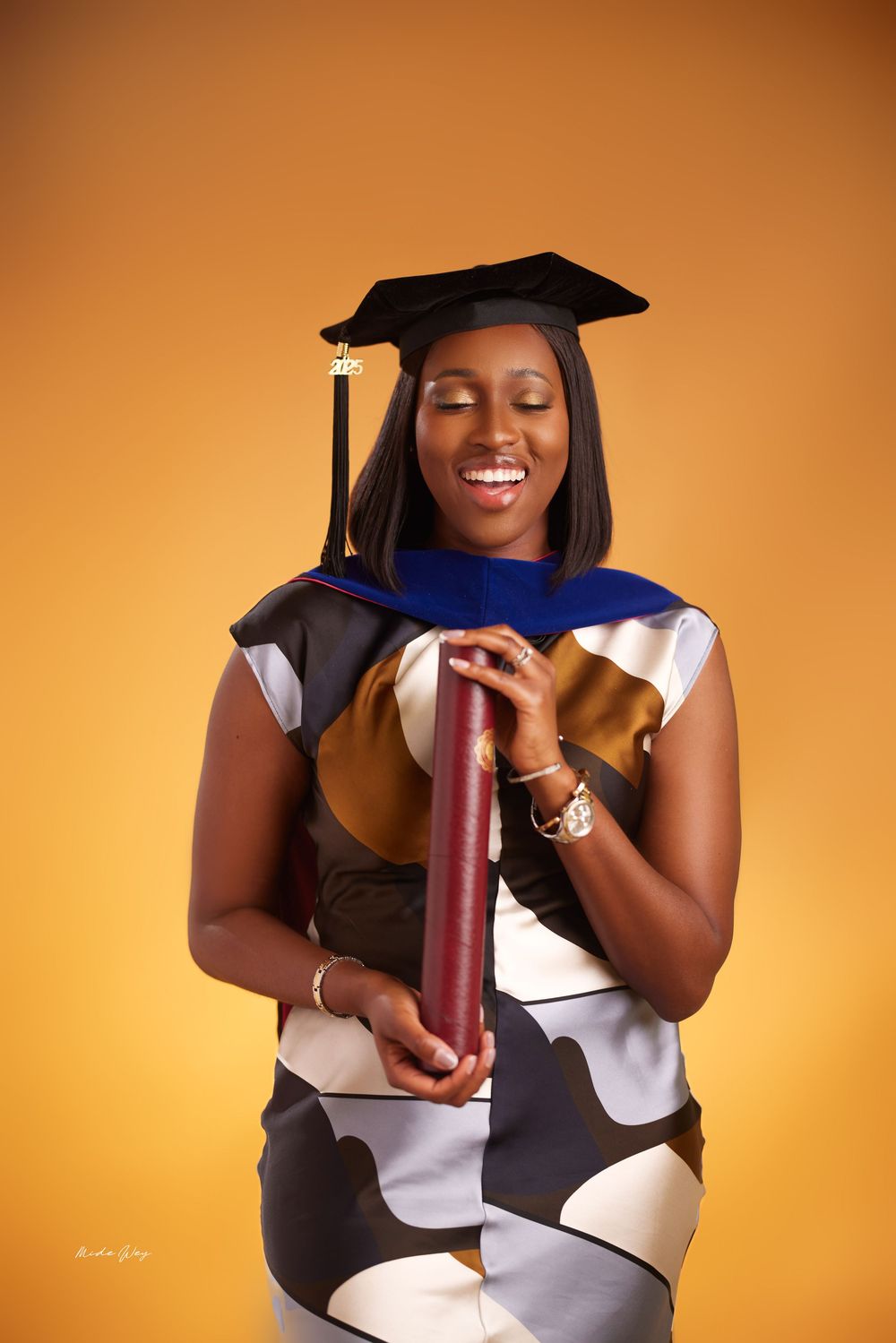 Graduate in academic regalia poses with diploma against warm orange background.