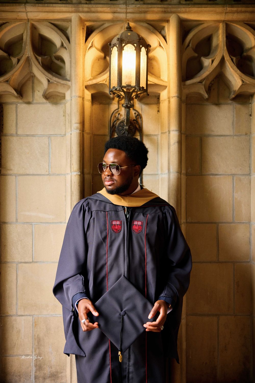 Graduate in academic regalia stands beneath gothic stone archway with ornate hanging lantern.