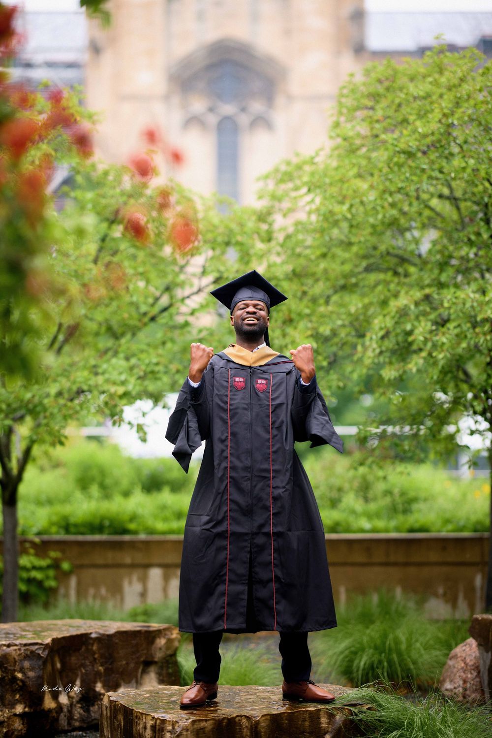 Graduate in academic regalia stands triumphantly on stone ledge surrounded by lush greenery and gothic architecture.