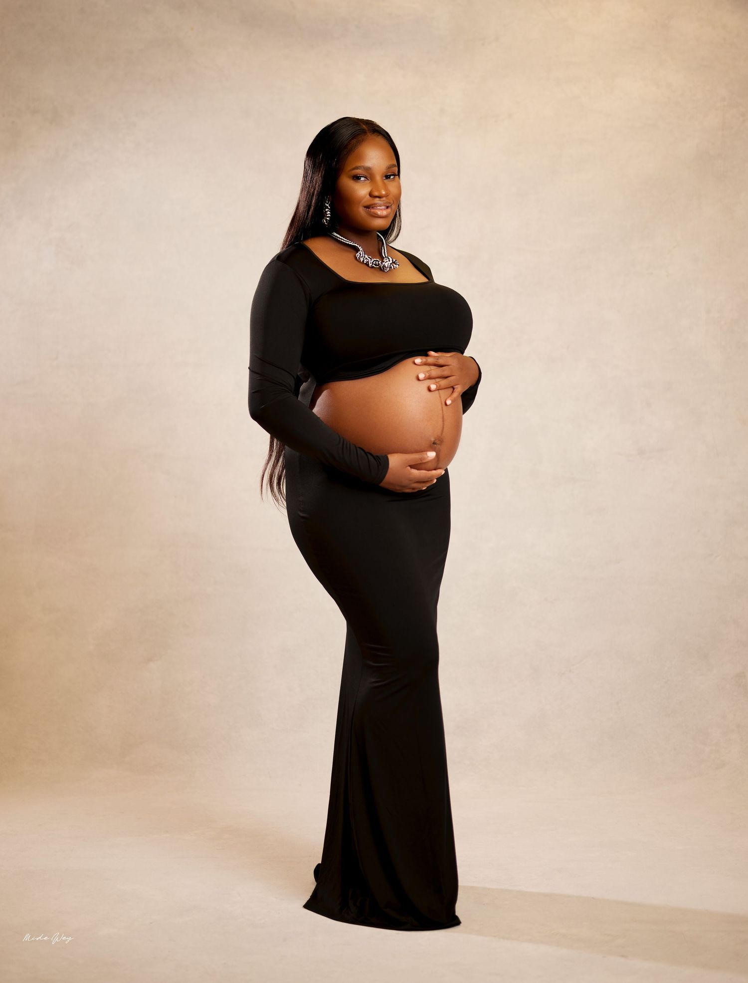 Expectant mother in a long black formal dress poses for a maternity photo against a neutral studio backdrop.