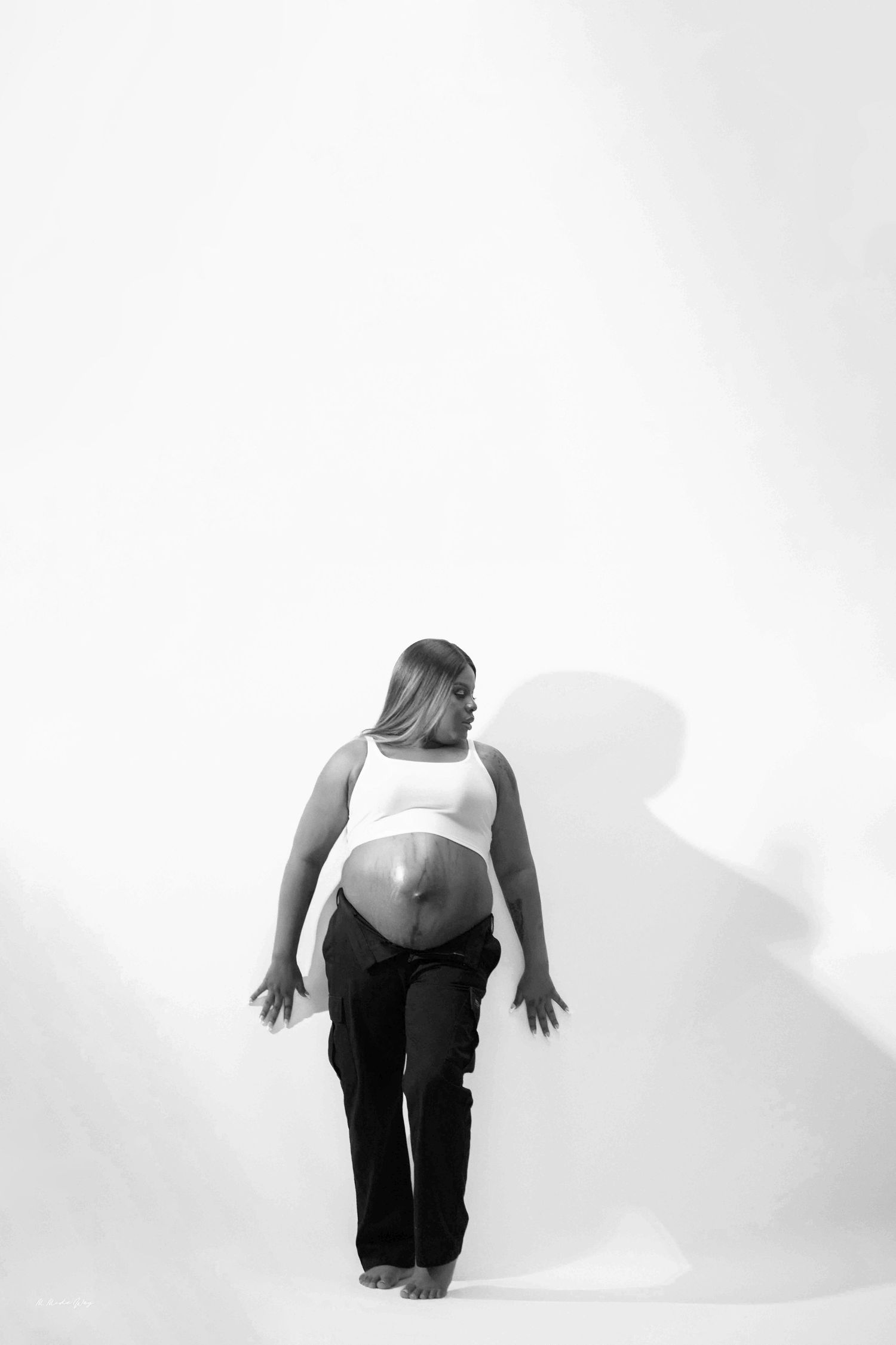 Black and white artistic portrait showing a person from behind wearing a white crop top and dark pants against a plain background.
