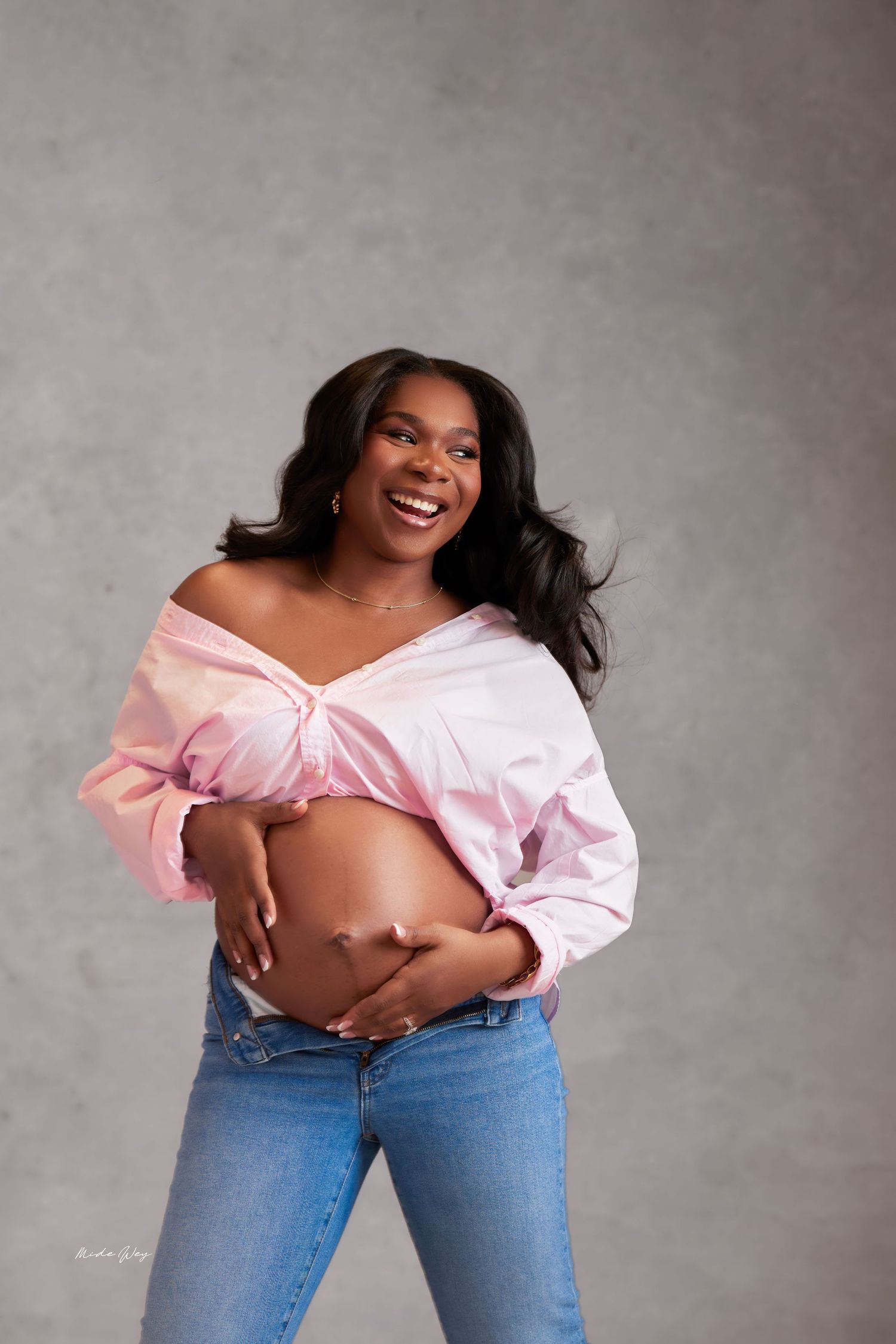A person in blue jeans and pink off-shoulder top poses playfully against a gray studio backdrop.