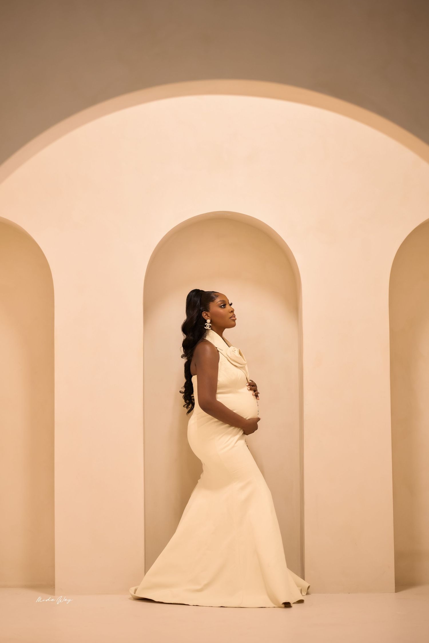 A woman in a flowing white dress poses gracefully against a minimalist cream-colored archway background.