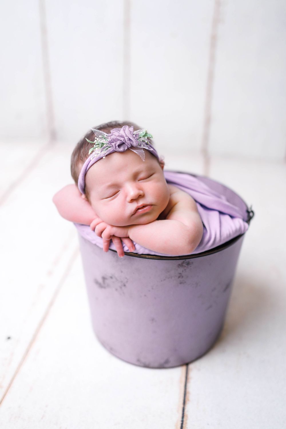 Sweet newborn sleeping peacefully in a purple bucket wearing a floral headband and wrapped in lavender fabric.  lawton oklahoma newborn