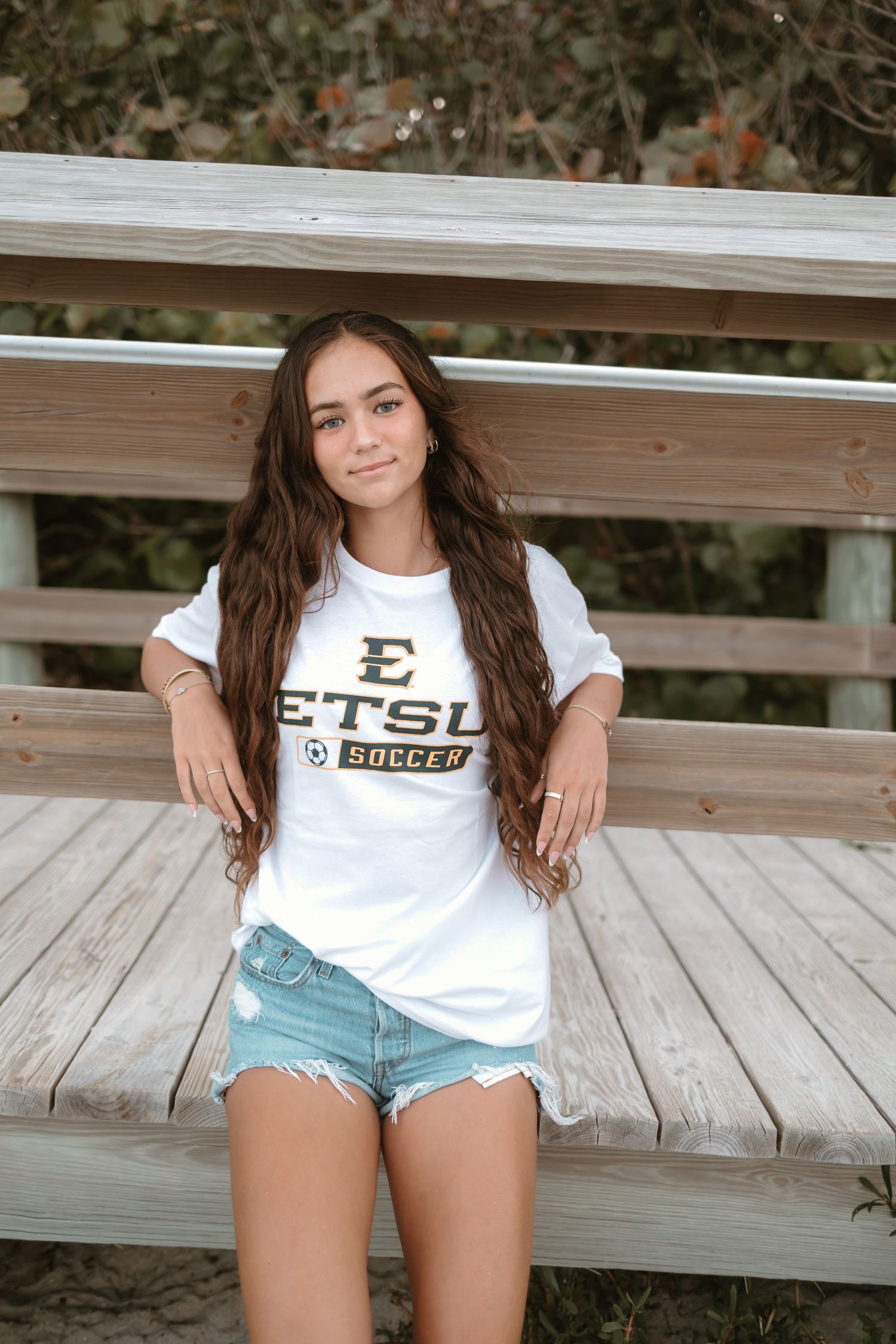 Person wearing ETSU Soccer t-shirt and denim shorts posing on wooden deck.